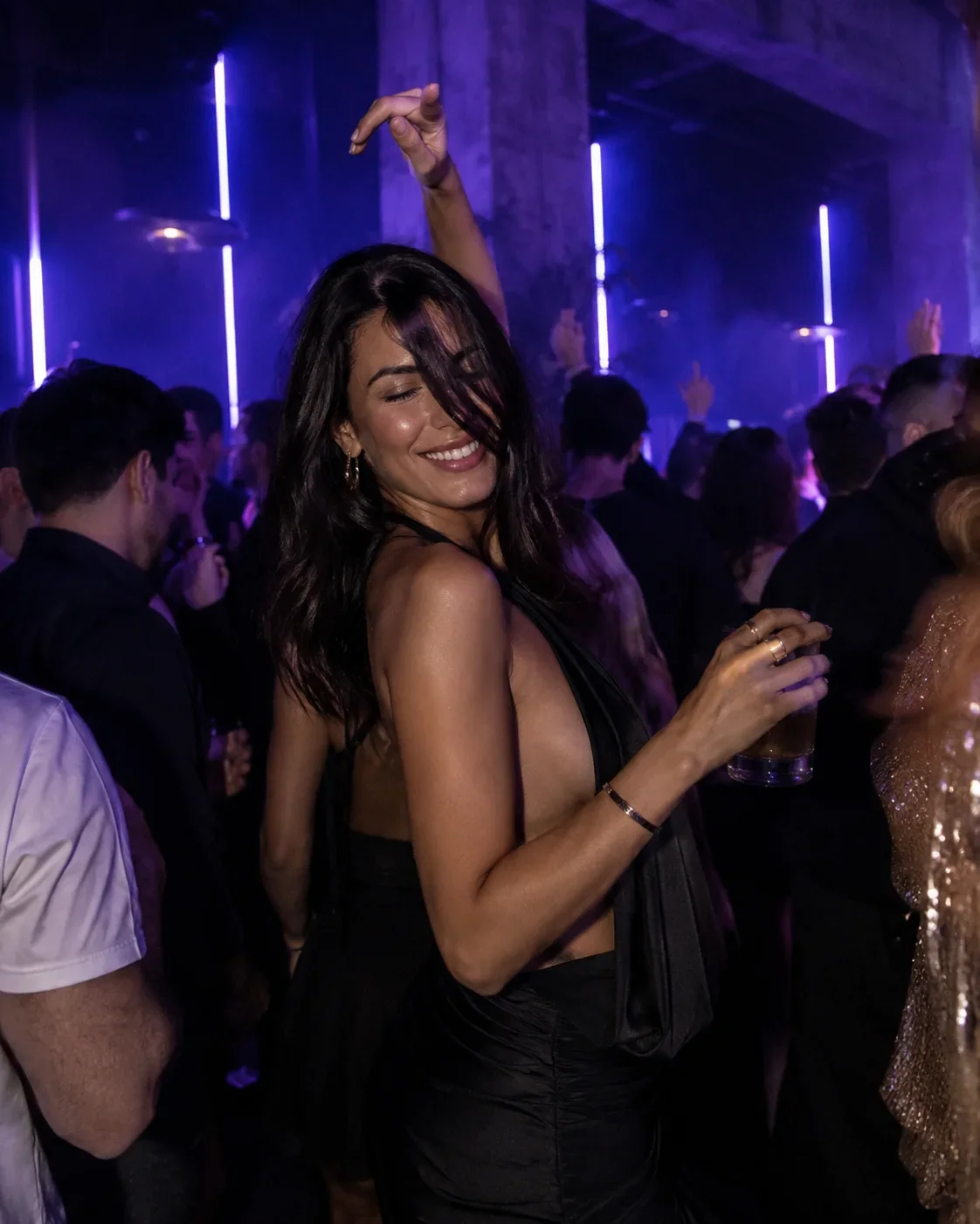 Elegante Frau im schwarzen Kleid tanzt mit einem Drink zwischen den Partygästen.